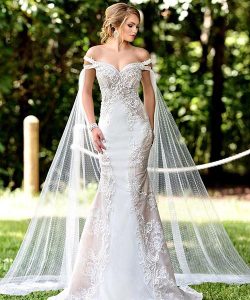 Woman in an off-the-shoulder, lace wedding gown with long, sheer cape stands outdoors on grass.