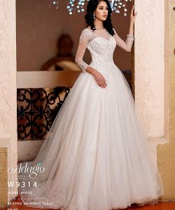 Woman wearing an ivory white beaded shimmer tulle ball gown with sheer sleeves, standing indoors by an archway.