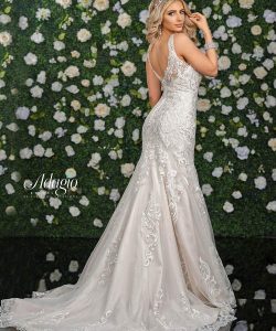 Woman in a white lace wedding gown stands in front of a green floral wall, looking over her shoulder.