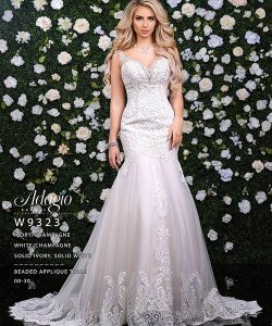 Woman in a beaded ivory wedding gown stands in front of a flower-covered wall, looking directly at the camera.