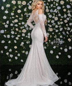 A woman in a long, white, lace gown with off-the-shoulder sleeves stands in front of a flower-covered wall.