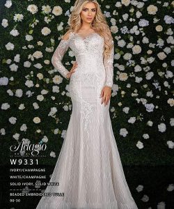 A woman in a fitted, off-shoulder, embroidered ivory gown stands in front of a flower-covered wall.