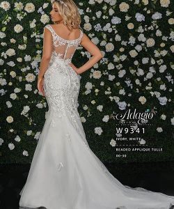 A woman in a white, beaded applique tulle wedding dress stands with her back to a flower-covered wall.