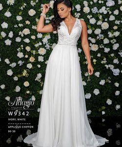 Woman wearing a sleeveless white applique chiffon gown stands in front of a green wall decorated with white flowers.