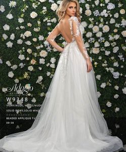 Woman in an open-back, long-sleeve white bridal gown stands before a flower-covered wall, facing sideways.