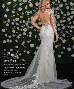 Woman in an open-back, long-sleeve, ivory lace wedding gown stands before a wall covered with white and green flowers.