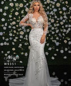 Woman wearing a long-sleeve, lace-embellished white wedding gown stands in front of a floral wall.