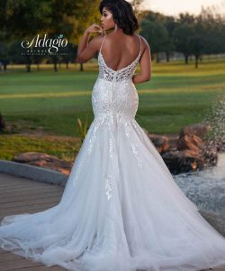A woman in a white, backless, lace mermaid wedding gown stands outdoors near grass and water.