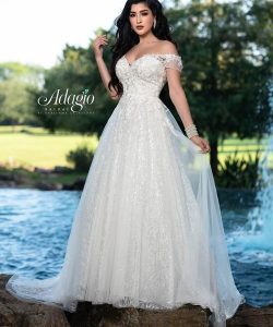 A woman in a white, off-the-shoulder, lace wedding dress stands outdoors near a fountain and greenery.