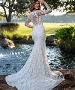 A woman in a long-sleeved, lace wedding gown stands outdoors by a pond, facing away from the camera.
