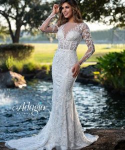 Woman wearing a long-sleeved, lace wedding gown stands outdoors by a pond with trees in the background.