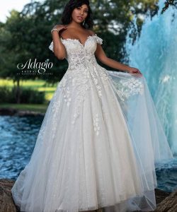 A woman in a white off-the-shoulder, lace-embellished wedding dress stands outdoors near a fountain and trees.