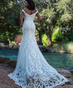 A woman stands outdoors by water, wearing a white, lace off-the-shoulder wedding gown with a long train.