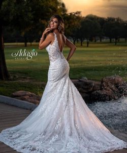A woman in a white lace wedding gown stands outdoors on a wooden path near grass, trees, and a water feature.