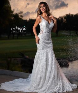 A woman wearing a white lace wedding dress stands outdoors near a pond at sunset.