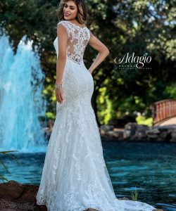 A woman in a lace wedding dress stands by a pond with a fountain and trees in the background.