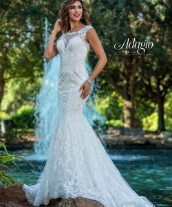 A woman in a sleeveless, lace mermaid wedding gown stands outdoors near a fountain and greenery.