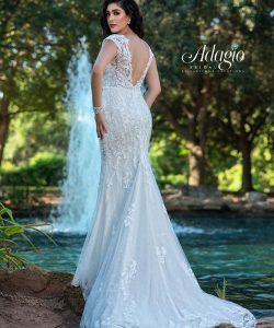Woman in a white lace wedding gown stands by a fountain and trees, looking over her shoulder.