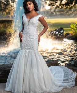 Woman in a fitted white lace mermaid wedding gown stands outdoors near a fountain, with greenery in the background.