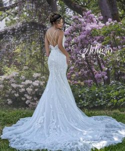 A woman in a lace-back white bridal gown stands outdoors among blooming purple and white flowers and green trees.