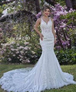 A woman in a white lace wedding dress stands on grass in front of blooming pink and white flowers.