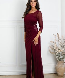 Woman in a burgundy evening gown with sheer sleeves stands indoors beside a vase of dried flowers against a white wall.